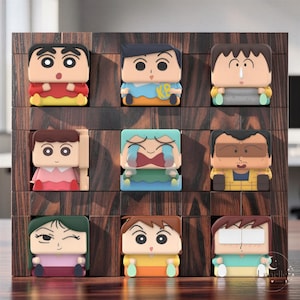 Puede incluir: Un estante de rejilla de madera oscura que contiene nueve figuras de personajes de dibujos animados de forma cuadrada. Cada figura presenta una expresión facial distinta, incluyendo sorpresa, llanto y neutralidad, con camisas y pantalones coloridos.