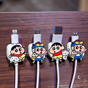 May include: Four white charging cables featuring decorative cable protectors shaped like the cartoon character Shin-chan. Each protector displays the character in various poses, some accompanied by a small white dog, attached to different USB connectors.