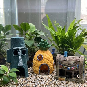 Könnte beinhalten: Drei Aquariendekorationen bestehend aus einem dunkelgrauen steinartigen Kopf, einem gelben Ananashaus mit grünen Blättern und einem braunen Gebäude in Holzoptik mit blauem Schornstein und einer Flaggenwimpelkette auf Kiesgrund.