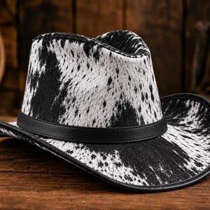 Op de afbeelding: Een cowboyhoed met een zwart-wit koeienprintpatroon over de kroon en de rand. De hoed is voorzien van een effen zwarte band rond de basis van de kroon en een zwarte bies langs de rand van de gebogen hoedrand voor een opvallend contrast.