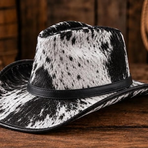 Op de afbeelding: Een cowboyhoed met een zwart-wit koeienhuidpatroon en een textuur die op bont lijkt. De hoed is voorzien van een gladde zwarte band rond de basis van de kroon en een bijpassende zwarte rand langs de brede rand van de hoed.