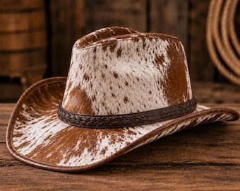 Op de afbeelding: Een cowboyhoed met een bruin en wit koeienhuidpatroon op de kroon en de rand. De hoed is voorzien van een donkerbruine gevlochten leren band rond de basis van de kroon en een bijpassende donkerbruine afwerking langs de rand van de hoed.