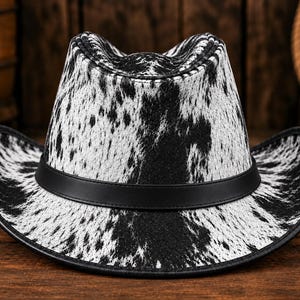 Op de afbeelding: Een cowboyhoed met een zwart-wit koeienprintpatroon over de kroon en de rand. De hoed is voorzien van een gladde zwarte band rond de basis van de kroon en een zwarte bies langs de rand van de gebogen hoedrand voor een duidelijk contrast.