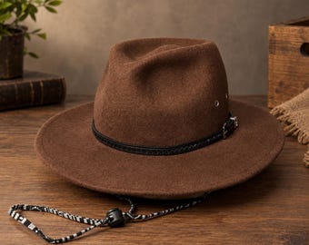 Op de afbeelding: Een bruine vilten fedora hoed met een zwarte gevlochten leren band en een zilveren gesp. De hoed is voorzien van metalen oogjes aan de zijkanten en een zwart-wit patroon kinband met een verstelbare plastic schuifsluiting voor de pasvorm.