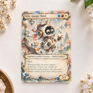 Può includere: Una carta collezionabile intitolata Kilo, Apogee Mind, che raffigura un robot bianco con cuffie in un paesaggio in stile acquerello. Il testo della carta include Legendary Artifact Creature, Robot Artificer, Haste e le regole per l'abilità proliferare, con una forza e costituzione di 3/3.