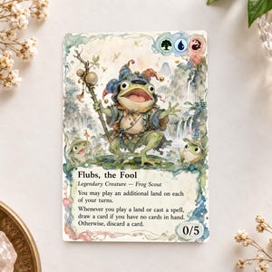May include: A rectangular trading card titled Flubs, the Fool, featuring a watercolour illustration of a frog wearing a jester hat and holding a staff. The card displays game text, mana symbols, and a power and toughness value of 0/5.