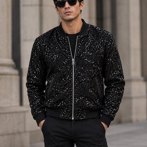 Könnte beinhalten: Eine schwarze Bomberjacke, die mit kleinen, reflektierenden Pailletten besetzt ist. Die Jacke verfügt über einen silbernen Reißverschluss in der Mitte, einen schwarzen Rippstrickkragen, schwarze Rippstrickbündchen und zwei Seitentaschen mit schwarzer Einfassung.
