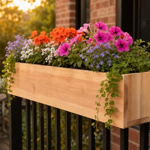 May include: A rectangular light-colored wooden planter box mounted on a black railing, filled with a vibrant arrangement of bright pink petunias, orange geraniums, small white flowers, and trailing green foliage that spills over the edges.