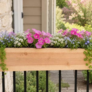 May include: A rectangular light brown wooden planter box filled with a variety of vibrant flowers, including bright pink petunias, small white alyssum, and light blue lobelia, with green trailing vines cascading over the sides of the container.