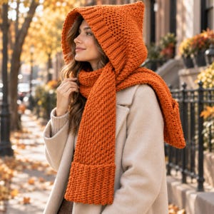 May include: A bright orange hooded scarf featuring a thick, textured knit pattern with a ribbed border along the hood and the scarf ends. The accessory is worn over a light beige coat, providing full coverage for the head and neck area.