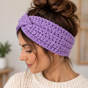May include: A lavender crochet headband featuring a textured stitch pattern and a central twisted knot detail. The accessory is crafted from yarn and designed to be worn around the head, providing a wide band that covers the ears and forehead area.