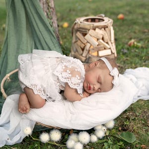 May include: A realistic baby doll with closed eyes, wearing a white lace-trimmed romper and a matching white headband with a small bow. The doll is lying on a white fabric cushion, positioned within a woven basket, and appears to be sleeping.