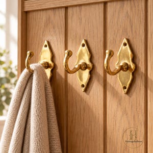 May include: Three polished brass hooks with diamond-shaped backplates mounted on a vertical oak wood panel. A beige knit garment hangs from the leftmost hook. The bottom right corner features a circular logo with the text HeritageByBrass.