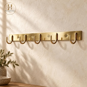 May include: A horizontal brass wall rack featuring five U-shaped hooks attached to a rectangular mounting plate. The metal has a brushed finish and is secured with three visible screws. The logo HeritageByBrass is printed in the top left corner.