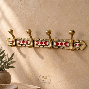 May include: A horizontal brass wall hook rack featuring four curved hooks. The rectangular base plate is decorated with inlaid white and red diamond-shaped tiles arranged in a repeating pattern. The metal has a warm, aged finish with visible screws.