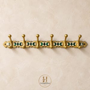 Op de afbeelding: Een horizontale wandkapstok van messing met zes haken met bolvormige uiteinden. De centrale balk is versierd met afwisselende geometrische inlegwerk van donkergroene en witte steen. Het logo HeritageByBrass is onderaan in het midden zichtbaar.