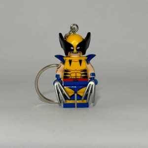 May include: A keyring featuring a yellow and blue plastic figure with a black mask and pointed ears. The figure has silver-coloured claws extending from each hand, a yellow torso with black stripes, and a blue belt with a red square buckle.