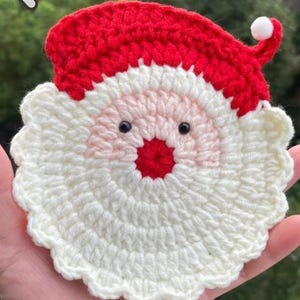 Peut inclure: Un dessous de verre circulaire au crochet en forme de visage de Père Noël. Le motif présente un bonnet rouge avec un pompon blanc, un visage rose pâle, un nez rouge vif et deux petits yeux en perles noires. La partie inférieure est une barbe blanche texturée.