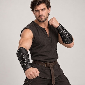 May include: A pair of black leather forearm bracers featuring three horizontal straps with silver-toned metal buckles and circular rivets along the edges. The bracers are worn on the forearms, paired with a dark gray sleeveless tunic and a brown belt.