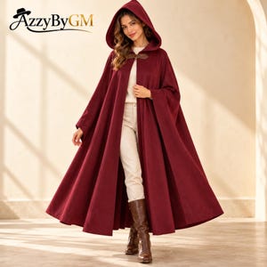 May include: A floor-length deep burgundy hooded cloak featuring a structured hood and a single brown leather-style tab closure at the neck. The garment is worn over a white long-sleeve top, light-colored trousers, and brown leather boots.