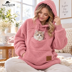 Könnte beinhalten: Ein altrosa Kapuzenpullover mit langen Ärmeln aus flauschigem, strukturiertem Fleece-Material. Das Kleidungsstück verfügt über eine große Bauchtasche, in der ein kleines, flauschiges, weiß-braunes Kätzchen sitzt. Ein brauner Aufnäher ist auf der Tasche.