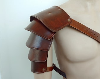 Può includere: Armatura per spalla in pelle marrone con più strati e una cinghia sul petto.