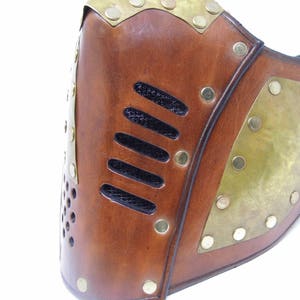 May include: A brown leather mask with brass accents and a black mesh panel. The mask has a steampunk aesthetic.
