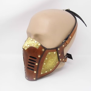 May include: A brown leather mask with brass accents and a strap. The mask has a geometric design with a series of small holes in the front.
