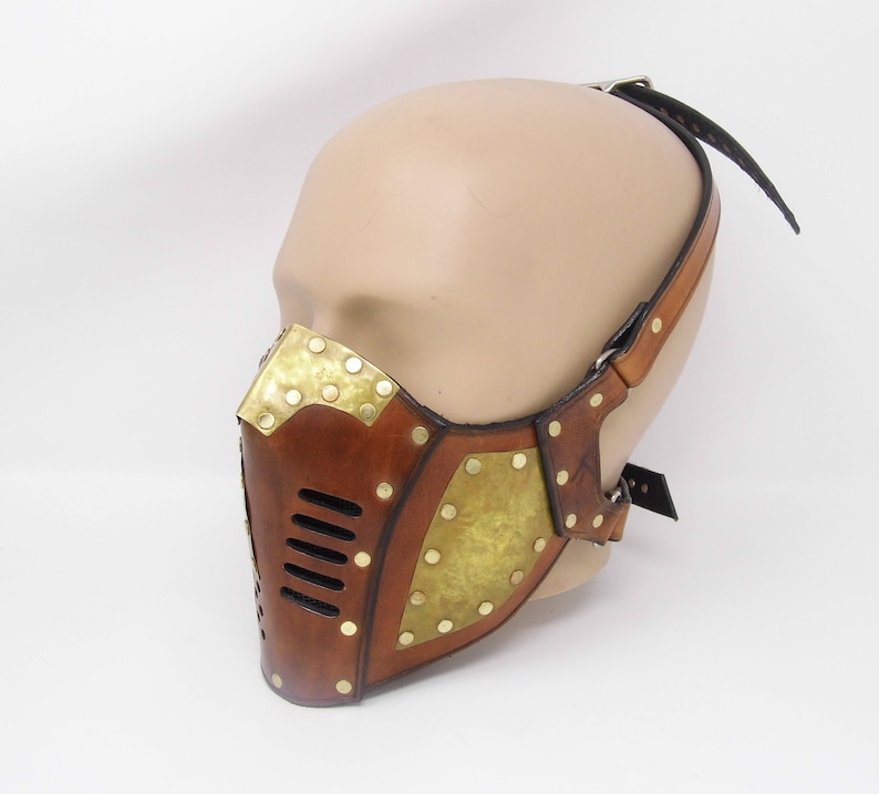 May include: A brown leather mask with brass accents and a strap. The mask has a geometric design with a series of small holes in the front.