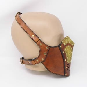 May include: A brown leather mask with brass accents and a perforated metal panel. The mask is attached to a brown leather strap that goes over the head.