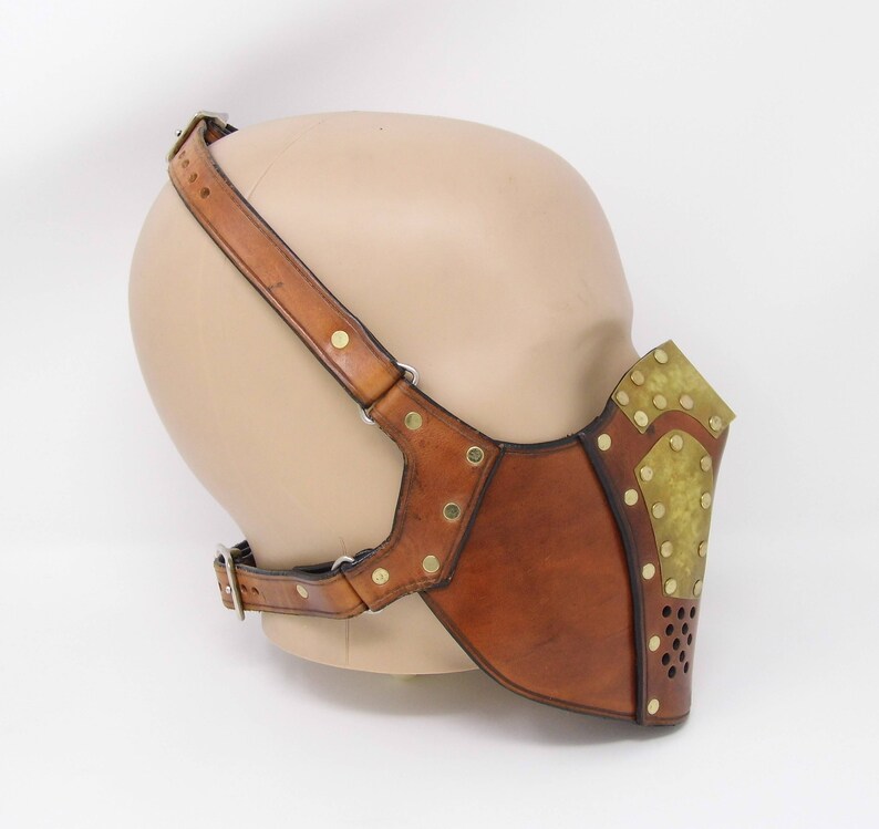 May include: A brown leather mask with brass accents and a perforated metal panel. The mask is attached to a brown leather strap that goes over the head.