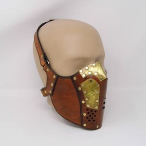 May include: A brown leather face mask with brass accents and rivets. The mask has a geometric design and is secured with a strap that goes around the head.