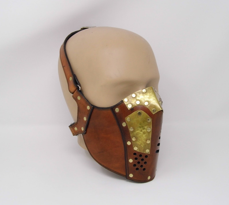 May include: A brown leather face mask with brass accents and rivets. The mask has a geometric design and is secured with a strap that goes around the head.