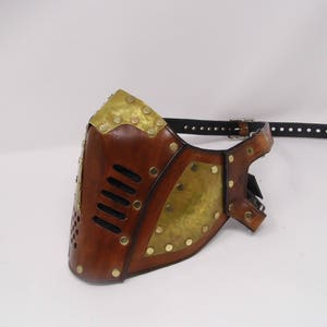 May include: A brown leather mask with brass accents and a black leather strap. The mask has a geometric design with a series of small holes.