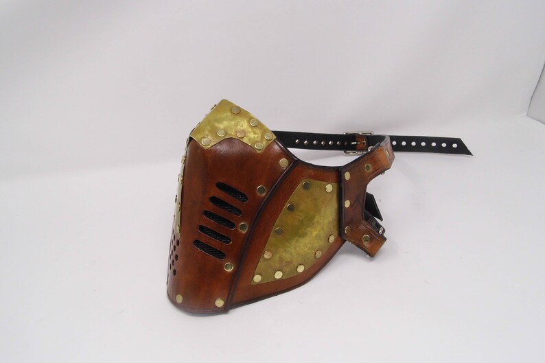 May include: A brown leather mask with brass accents and a black leather strap. The mask has a geometric design with a series of small holes.
