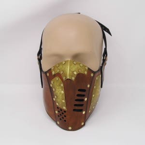 May include: A brown leather mask with brass accents and a metal mesh grille. The mask has a strap that goes over the head.