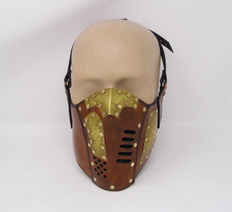 May include: A brown leather mask with brass accents and a metal mesh grille. The mask has a strap that goes over the head.