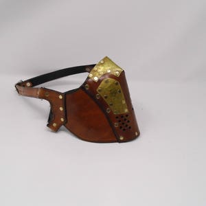 May include: A brown leather face mask with brass accents and a strap. The mask has a perforated design and is secured with a strap.