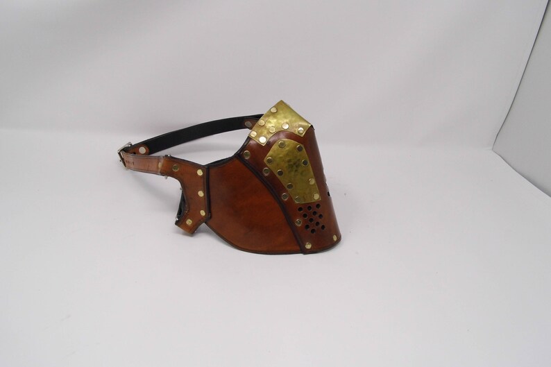May include: A brown leather face mask with brass accents and a strap. The mask has a perforated design and is secured with a strap.