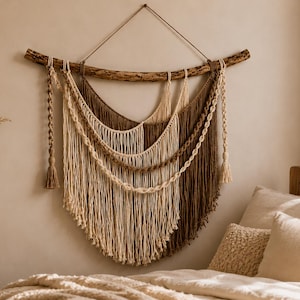 Puede incluir: Un tapiz de pared hecho de cuerda de macramé color crema y marrón suspendido de una rama de madera natural. La pieza presenta hebras de cuerda drapeadas en capas de colores alternos, con acentos laterales trenzados y borlas.