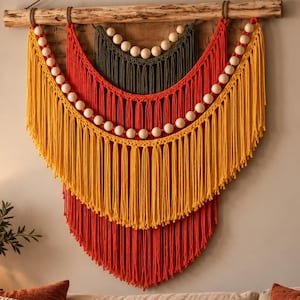 Puede incluir: Un tapiz de pared con cuerdas de macramé superpuestas en tonos verde oliva, naranja quemado y amarillo mostaza. El diseño incluye tres secciones curvas distintas adornadas con cuentas de madera natural, suspendidas de una rama de madera.