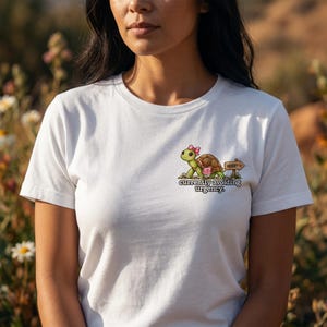 Op de afbeelding: Een wit T-shirt met korte mouwen en een afbeelding van een groene schildpad met een roze strik en een klein roze tasje. Naast de schildpad staat een houten bordje met de tekst HURRY. Onder de afbeelding staat de tekst currently avoiding urgency.