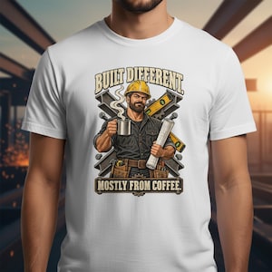 Pode incluir: Uma t-shirt branca com um gráfico de um trabalhador da construção civil com capacete amarelo, segurando uma caneca fumegante e uma planta. O texto diz BUILT DIFFERENT. MOSTLY FROM COFFEE. O design inclui vigas de aço e um nível.