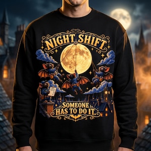Könnte beinhalten: Ein schwarzes Sweatshirt mit Rundhalsausschnitt und einem Grafikdruck von drei Fledermäusen in blauen Uniformen, die an einem Vollmond arbeiten. Der Text lautet NIGHT SHIFT und SOMEONE HAS TO DO IT, ergänzt durch eine Stadtkulisse.