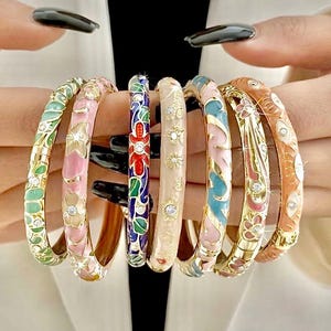 May include: A collection of seven hinged metal bangles featuring colorful enamel patterns, including floral, leaf, and sun motifs, accented with small embedded clear crystals. Each bracelet has a polished gold-tone finish and a secure clasp mechanism.