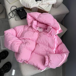 May include: A bright pink puffer jacket featuring a hood, a front zip fastening, and long sleeves with elasticated cuffs. The jacket has a quilted, padded construction with a high collar and a label visible on the inside of the neckline.
