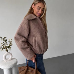 May include: A taupe-colored, textured fleece jacket with a collared neckline, long sleeves, and a zippered front closure, paired with dark blue denim jeans and a brown suede handbag featuring a buckle detail and dual top handles.