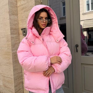 Peut inclure: Une doudoune rose vif dotée d'une grande capuche, d'un col montant et de manches longues avec des poignets élastiqués. La veste présente une fermeture éclair sur le devant et un design matelassé. Elle est portée avec un jean bleu clair.