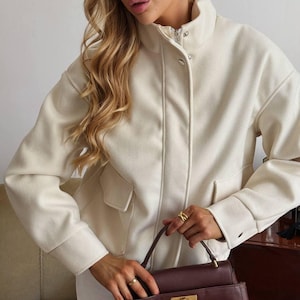 May include: A cream-colored jacket with a high collar, long sleeves, and two front flap pockets. The jacket features a front zipper closure concealed by a placket with snap buttons. A dark brown structured handbag with a gold-tone metal clasp is held.