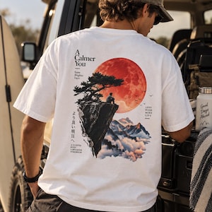 Könnte beinhalten: Ein weißes T-Shirt mit einem Grafikdruck auf der Rückseite. Das Motiv zeigt eine Person, die auf einer Klippe unter einem großen roten Mond sitzt, mit einem Baum, Bergen, Wolken und Vögeln. Der Text umfasst A Calmer You, Same Brighter Days, Less Noise More Life und japanische Schriftzeichen.